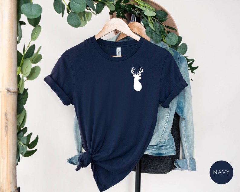 May include: A navy blue t-shirt with a white silhouette of a deer head with antlers on the front. The shirt is folded and hanging on a wooden hanger.