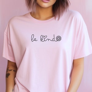Puede incluir: Una camiseta rosa con el texto "be kind" en letra negra con un peque&ntilde;o coraz&oacute;n y un gr&aacute;fico de flor.