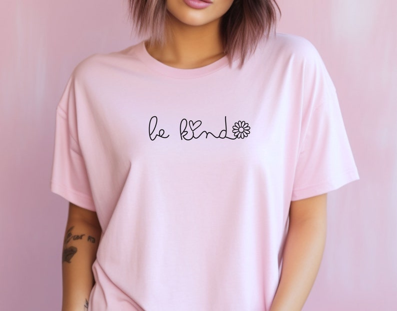 Puede incluir: Una camiseta rosa con el texto "be kind" en letra negra con un peque&ntilde;o coraz&oacute;n y un gr&aacute;fico de flor.