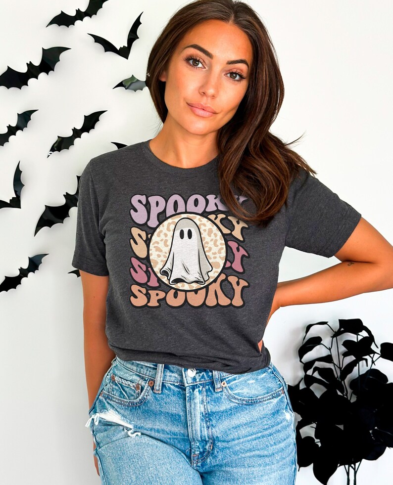 Puede incluir: Una camiseta gris oscuro con un gr&aacute;fico de un fantasma blanco en un c&iacute;rculo rosa, morado y marr&oacute;n con la palabra "SPOOKY" repetida alrededor del c&iacute;rculo.