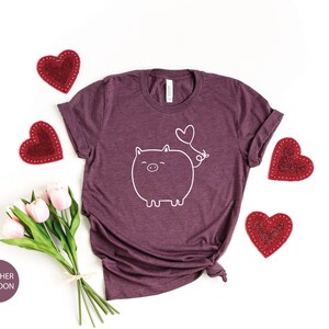 Peut inclure: Un t-shirt en marron chin&eacute; avec un contour blanc d'un cochon avec un ballon en forme de c&oelig;ur qui sort de son arri&egrave;re-train. Le t-shirt est entour&eacute; de c&oelig;urs rouges et d'un bouquet de tulipes roses.