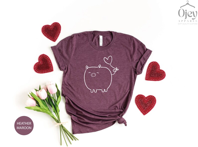 Peut inclure: Un t-shirt en marron chin&eacute; avec un contour blanc d'un cochon avec un ballon en forme de c&oelig;ur qui sort de son arri&egrave;re-train. Le t-shirt est entour&eacute; de c&oelig;urs rouges et d'un bouquet de tulipes roses.