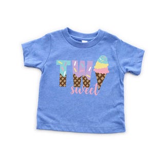 May include: Blue short-sleeved t-shirt with the text "TWO sweet" in pink and white. The number two is made of ice cream cones with sprinkles.