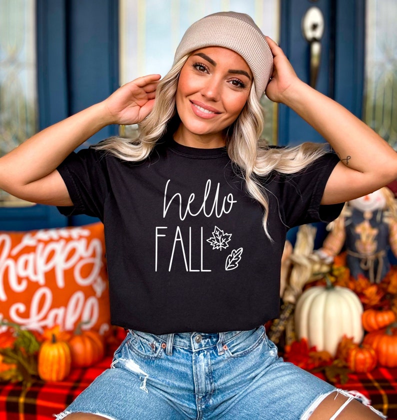 May include: A black t-shirt with the text "hello fall" in white lettering and a white outline of a leaf. The shirt is being worn by a person with blonde hair and a beige beanie.