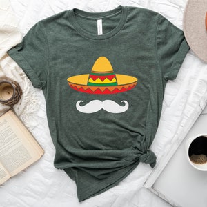 May include: A green t-shirt with a white mustache and a colorful sombrero on it. The shirt is on a white surface with a book, a clock, a cup of coffee, and a hat.