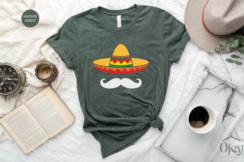 May include: A green t-shirt with a white mustache and a colorful sombrero on it. The shirt is on a white surface with a book, a clock, a cup of coffee, and a hat.