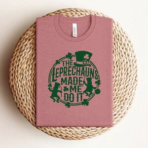 May include: A pink t-shirt with a green graphic that says "The Leprechauns Made Me Do It". The graphic features a leprechaun with a hat and a pot of gold.