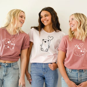 Peut inclure: Trois femmes portant des t-shirts roses et blancs avec de jolies illustrations d'animaux et des ballons en forme de c&oelig;ur. Les t-shirts repr&eacute;sentent un chien, un ours et une baleine.