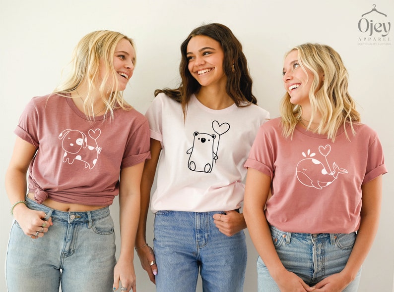 Peut inclure: Trois femmes portant des t-shirts roses et blancs avec de jolies illustrations d'animaux et des ballons en forme de c&oelig;ur. Les t-shirts repr&eacute;sentent un chien, un ours et une baleine.