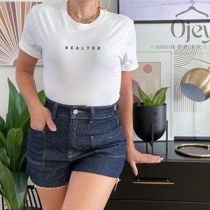 Op de afbeelding: Een wit T-shirt met het woord "REALTOR" erop gedrukt. De persoon draagt een blauwe denim short.