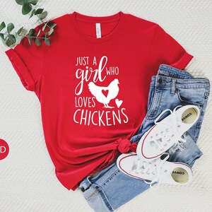 Puede incluir: Camiseta roja con texto blanco que dice "Just a girl who loves chickens" y una silueta blanca de una gallina con un coraz&oacute;n en el pecho.