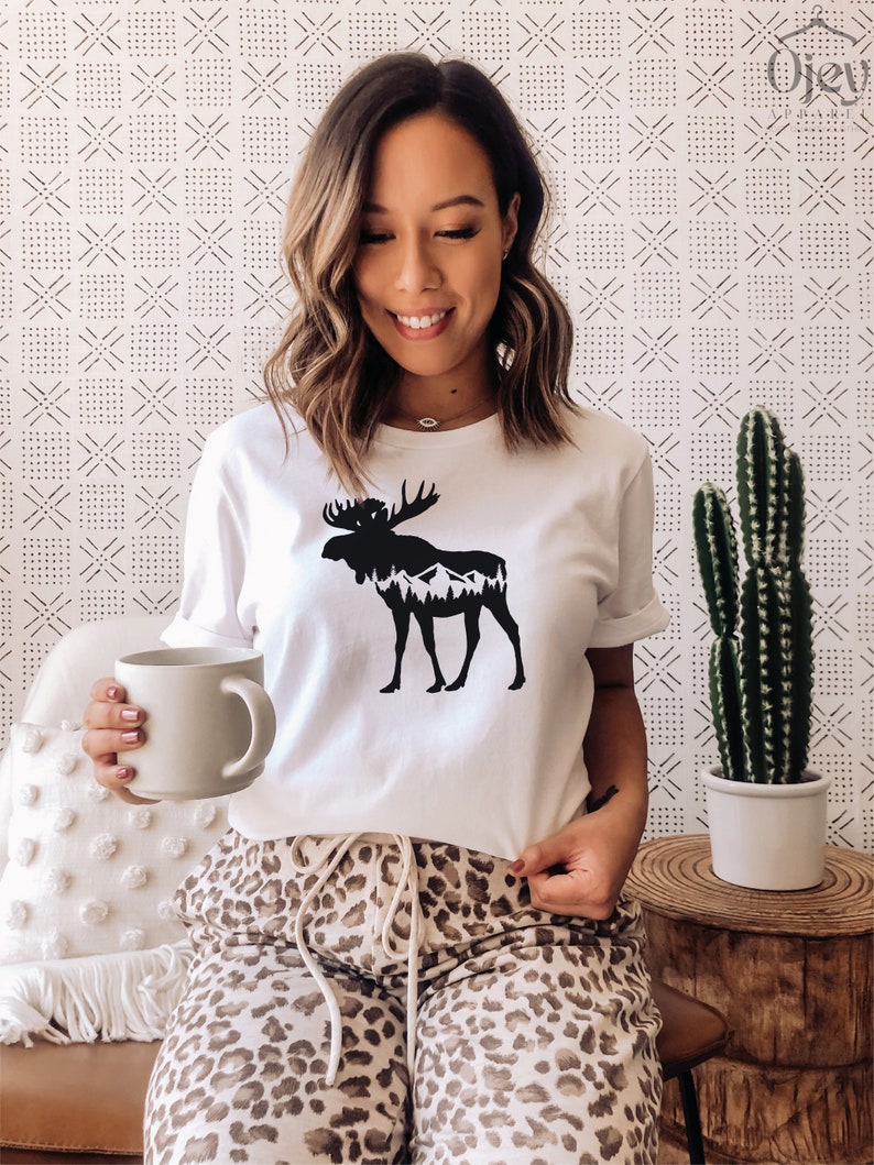 May include: White t-shirt featuring a black silhouette of a moose with mountains and trees inside. The model is wearing leopard print pants and holding a beige mug. A cactus in a white pot sits on a wooden side table.