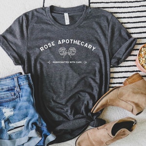 May include: Dark gray t-shirt with the text "ROSE APOTHECARY" and two floral illustrations. Below the text is "HANDCRAFTED WITH CARE". The shirt is paired with denim shorts and tan ankle boots. A small potted cactus is also in the image.
