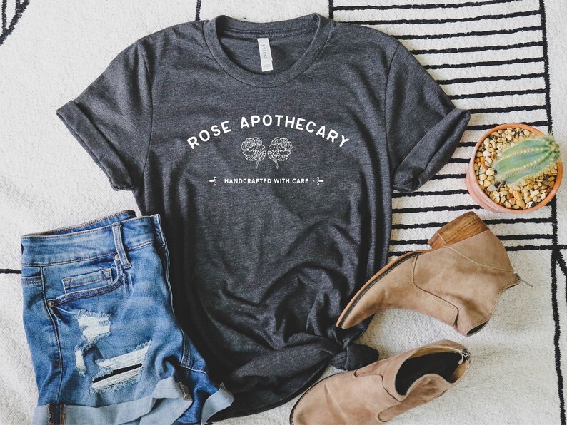 May include: Dark gray t-shirt with the text "ROSE APOTHECARY" and two floral illustrations. Below the text is "HANDCRAFTED WITH CARE". The shirt is paired with denim shorts and tan ankle boots. A small potted cactus is also in the image.