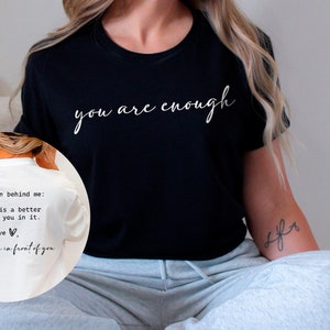 May include: A black t-shirt with the text "you are enough" in white script lettering. The back of the shirt has a message written in white text that says "Dear person behind me: The world is a better place with you in it. Lots of love, the person in front of you."