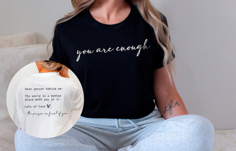 May include: A black t-shirt with the text "you are enough" in white script lettering. The back of the shirt has a message written in white text that says "Dear person behind me: The world is a better place with you in it. Lots of love, the person in front of you."