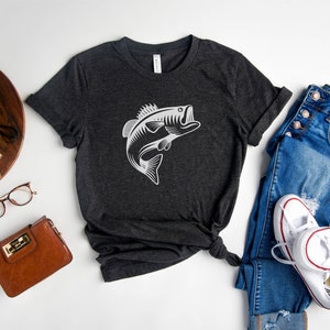 May include: Heather dark grey t-shirt featuring a white graphic of a bass fish leaping out of the water. The shirt is laid flat on a white surface with a pair of jeans, a brown purse, a camera, and a potted plant.