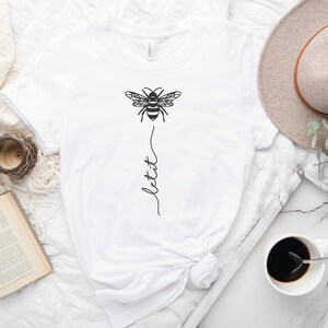 May include: White t-shirt with a black bee graphic and the text "let it" written vertically below the bee.