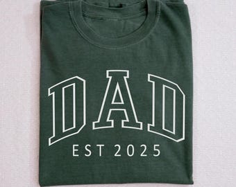 Puede incluir: Una camiseta verde oscuro con un contorno blanco de la palabra "DAD" en una fuente de estilo universitario. El texto "EST 2025" está debajo de la palabra "DAD".