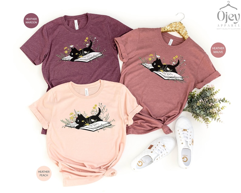 Puede incluir: Tres camisetas con un gato negro leyendo un libro rodeado de flores. Las camisetas son de color marr&oacute;n jaspeado, malva jaspeado y melocot&oacute;n jaspeado.
