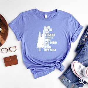 May include: A light blue t-shirt with a white graphic of a tree and the text "And into the forest I go to lose my mind and find my soul".