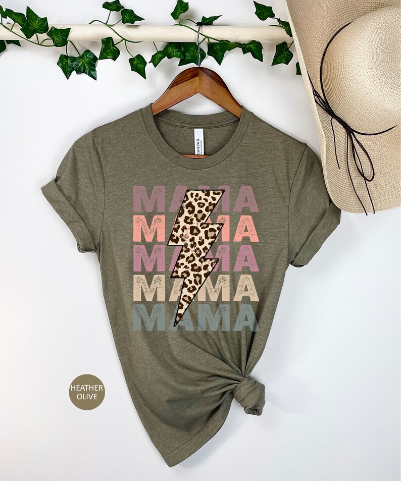 Op de afbeelding: Een groen T-shirt met het woord "MAMA" vier keer herhaald in een vervaagd regenboogkleurenschema. Een luipaardprint bliksemgrafiek is gecentreerd op het shirt.