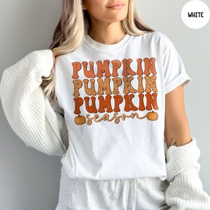 May include: White t-shirt with the words "PUMPKIN PUMPKIN PUMPKIN season" in orange and leopard print lettering. Two small orange pumpkins are on the bottom of the shirt. The shirt is a crew neck and has short sleeves.