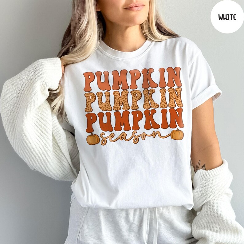 May include: White t-shirt with the words "PUMPKIN PUMPKIN PUMPKIN season" in orange and leopard print lettering. Two small orange pumpkins are on the bottom of the shirt. The shirt is a crew neck and has short sleeves.