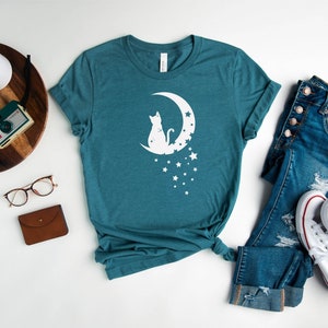 May include: A teal blue t-shirt with a white silhouette of a cat sitting on a crescent moon with stars. The t-shirt is laid flat on a white surface with a pair of glasses, a brown wallet, a wooden tray with a camera and a plant, and a pair of blue jeans and white sneakers.