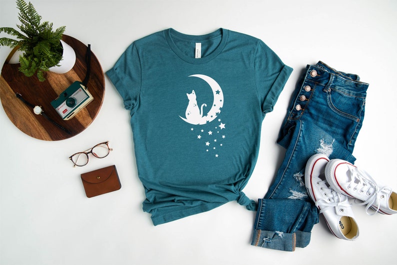 May include: A teal blue t-shirt with a white silhouette of a cat sitting on a crescent moon with stars. The t-shirt is laid flat on a white surface with a pair of glasses, a brown wallet, a wooden tray with a camera and a plant, and a pair of blue jeans and white sneakers.
