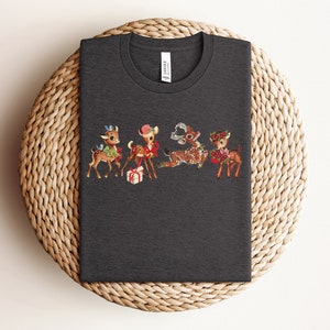 Pu&ograve; includere: Una maglietta grigia scura con un design natalizio vintage con quattro renne dei cartoni animati con accenti rossi e verdi. Le renne indossano cappelli e portano regali.