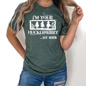 K&ouml;nnte beinhalten: Ein gr&uuml;nes T-Shirt mit einem wei&szlig;en Aufdruck, der "I'm your huckleberry... say when" lautet. Die Grafik zeigt drei Cowboys, die auf Pferden reiten.
