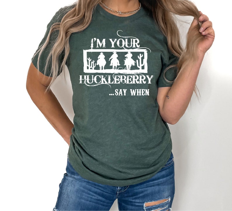K&ouml;nnte beinhalten: Ein gr&uuml;nes T-Shirt mit einem wei&szlig;en Aufdruck, der "I'm your huckleberry... say when" lautet. Die Grafik zeigt drei Cowboys, die auf Pferden reiten.