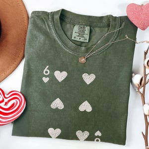 May include: Olive green t-shirt with a playing card design featuring the number 6 and white hearts. A gold necklace, a pink heart decoration, and a red and white heart-shaped lollipop are also visible.