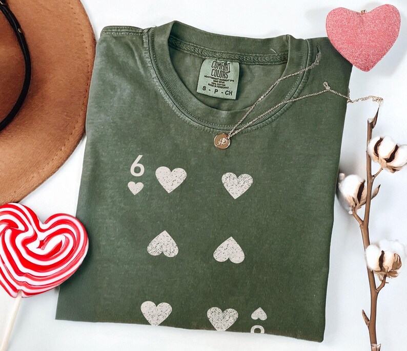 May include: Olive green t-shirt with a playing card design featuring the number 6 and white hearts. A gold necklace, a pink heart decoration, and a red and white heart-shaped lollipop are also visible.