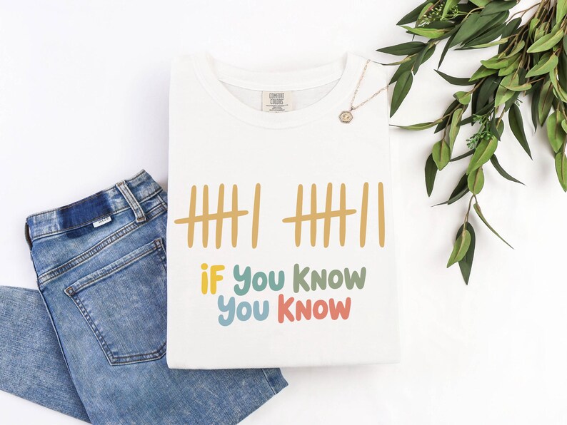 May include: White t-shirt with the text "If You Know You Know" in colorful letters. The shirt features a tally mark design in yellow. A pair of blue jeans is folded next to the shirt. A gold necklace and greenery are also in the image.