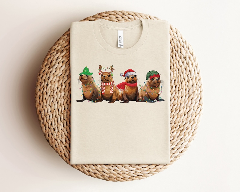 May include: A beige t-shirt with a Christmas-themed design featuring three sea lions wearing festive hats and lights. The sea lions are wearing a Santa hat, a reindeer headband, and a green elf hat.
