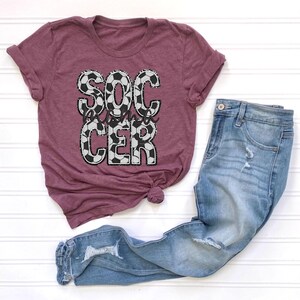 May include: A maroon t-shirt with the words "Soccer Mom" in white letters with a black and white soccer ball pattern.