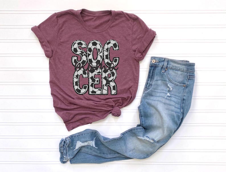 May include: A maroon t-shirt with the words "Soccer Mom" in white letters with a black and white soccer ball pattern.