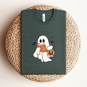 May include: A dark green t-shirt with a white ghost holding a book that says "Say Spooky". The ghost is carrying a small orange pumpkin with green stems and purple flowers.