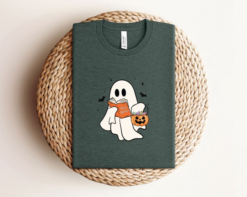 May include: A dark green t-shirt with a white ghost holding a book that says "Say Spooky". The ghost is carrying a small orange pumpkin with green stems and purple flowers.