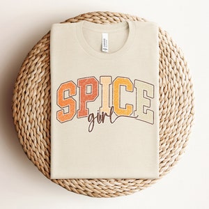 May include: A beige t-shirt with the text "SPICE girl" in a retro-style font with orange, yellow, and brown colors.