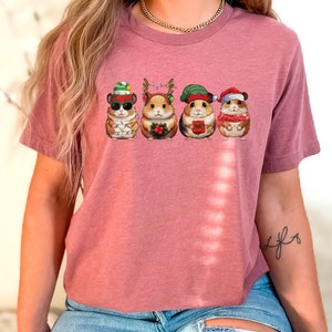 May include: A pink t-shirt with a graphic of five hamsters wearing Christmas hats and holding Christmas-themed items. The hamsters are wearing Santa hats, elf hats, reindeer antlers, and a stocking cap.