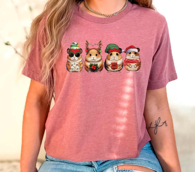 May include: A pink t-shirt with a graphic of five hamsters wearing Christmas hats and holding Christmas-themed items. The hamsters are wearing Santa hats, elf hats, reindeer antlers, and a stocking cap.