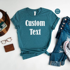 May include: A teal blue t-shirt with the text "Custom Text" in white. The shirt is laid flat on a white surface with a pair of jeans, a pair of white sneakers, a brown wallet, and a pair of glasses.