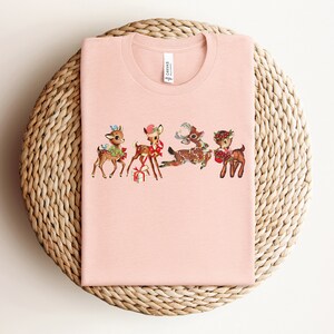 Pu&ograve; includere: Una maglietta rosa con un design natalizio vintage con quattro renne con fiocchi rossi e regali. Le renne sono in fila, rivolte a destra.