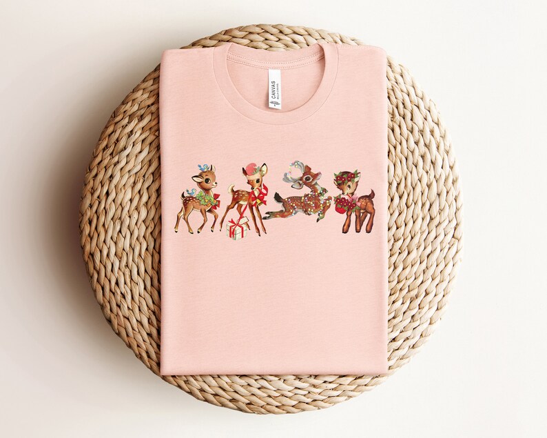 Pu&ograve; includere: Una maglietta rosa con un design natalizio vintage con quattro renne con fiocchi rossi e regali. Le renne sono in fila, rivolte a destra.