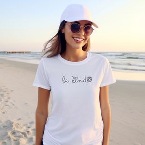 Puede incluir: Camiseta blanca con el texto "be kind" en cursiva y un peque&ntilde;o gr&aacute;fico de flor.
