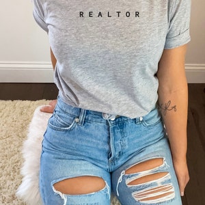 Op de afbeelding: Een grijs T-shirt met het woord "REALTOR" in zwart op de voorkant. De persoon die het shirt draagt, draagt ook een blauwe jeans met distressed details.