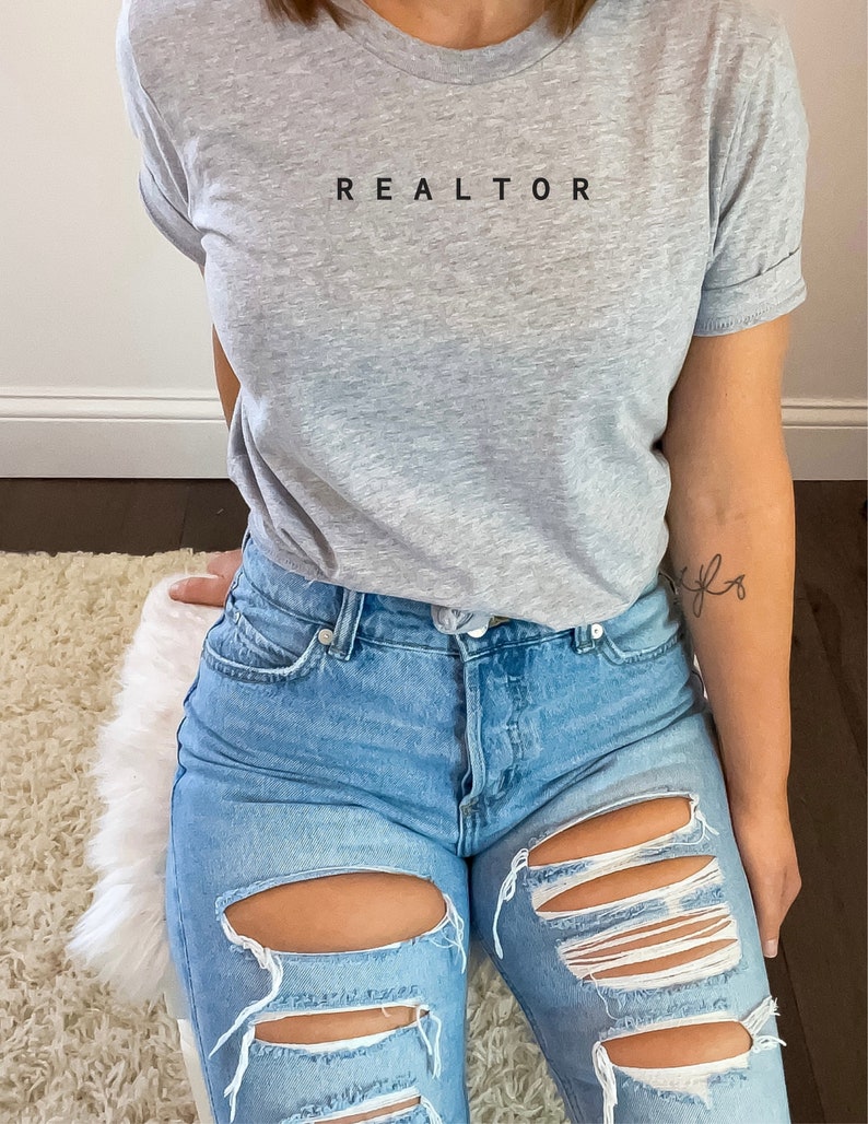 Op de afbeelding: Een grijs T-shirt met het woord "REALTOR" in zwart op de voorkant. De persoon die het shirt draagt, draagt ook een blauwe jeans met distressed details.